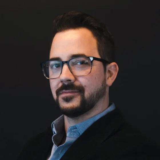 Head shot of a white man with black hair and trimmed beard, wearing glasses, a collared blue shirt, and a black suit jacket.