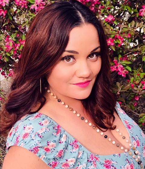 A Mexican American and Indigenous woman with dark hair streaked with red. She is wearing a floral blouse and long necklace and standing in front of a flowering bush