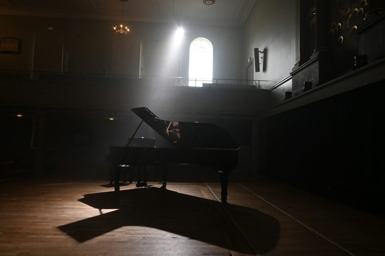 A black grand piano on a brown wooden floor as sun streams in from an arched-shape window above