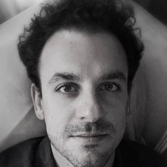 A black and white photo of a white man with tall dark hair. He's wearing a dark button-down shirt and in front of a lighter material