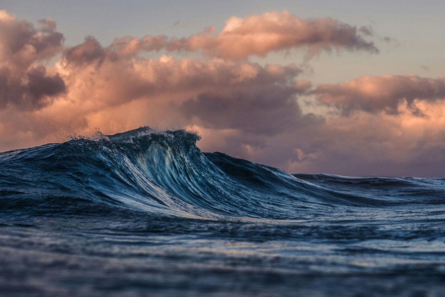 A wave mirrors the shape of the clouds above it, which seem to form a pink shelf over the water.