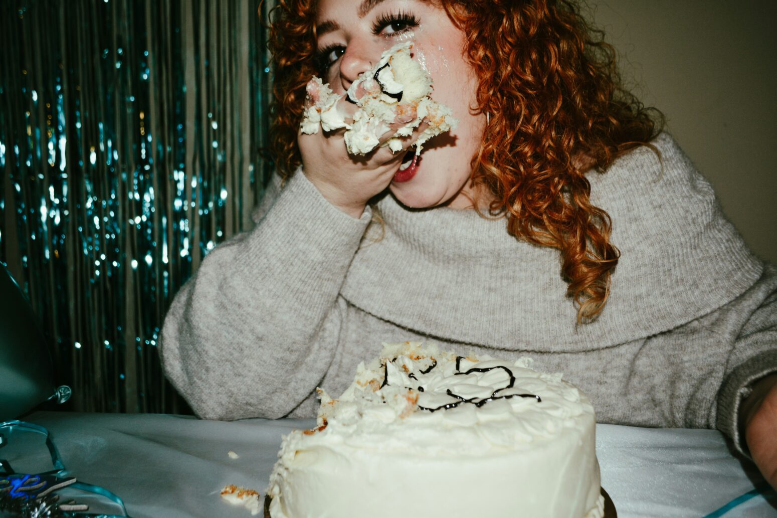 A white woman with red curly hair wearing a fuzzy white shawl-collared sweater sits in front of a white cake and eats out of her hands. Behind her, a blue streamer curtain shimmers.