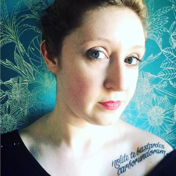 A white woman whose face is partly in shadow. She's wearing a V neck shirt and standing near floral blue wallpaper. She has a tattoo on her left shoulder that reads, "nolite te bastardes carborundorum"