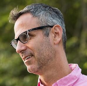 A side view of a white man with gray hair and glasses. He's wearing a pink checked button-down shirt and is outside