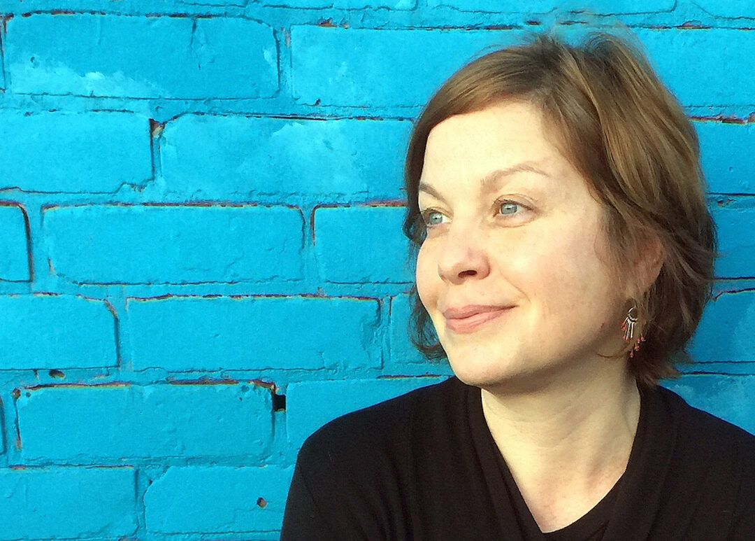 Sonja Livingston, a white woman with short brown hair, smiles in front of a brick wall painted blue. She looks to her right, at something in the middle distance. Her eyes match the color of the wall.