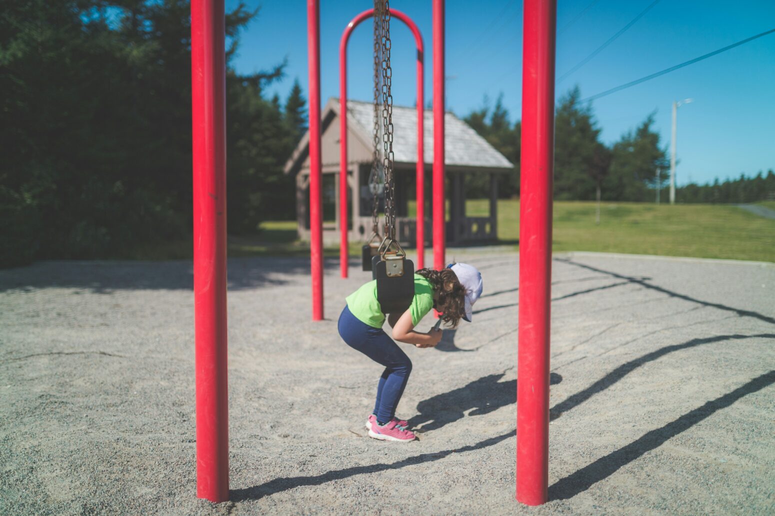 A child with long brown hair wearing a lime green tee shirt, jeans, and a baseball cap hangs from a swing on a red swing set in a park setting.