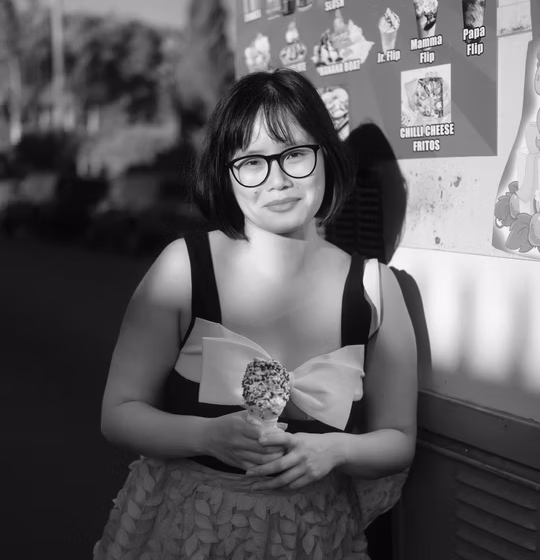 Black and white photo of an Asian person with short hair and big glasses. She is smiling and wearing a sleeveless shirt while standing in front of a bulletin board