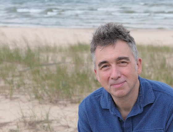 A white man with gray hair. He's wearing a textured blue shirt, and beach sand and grasses and then a body of water are behind him.