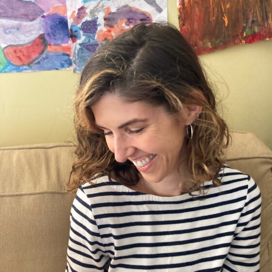 A white woman with brown hair to her shoulders, wearing a black and white striped shirt. She's smiling and looking to the side and down, as she sits in front of a tan couch with colored images on papers behind her.