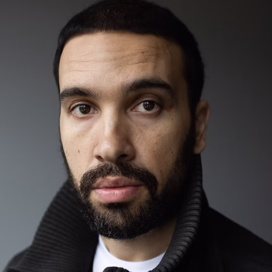 A Black man with short hair and a beard. He's wearing a black jacket with tall collar and a white shirt underneath, and in front of a gray background