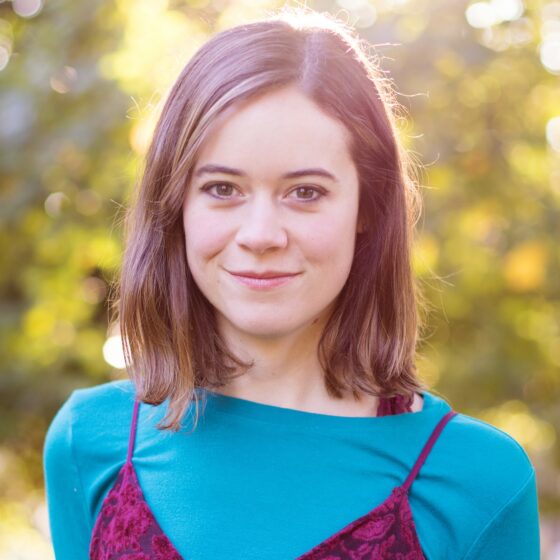 A white woman with brown hair to her shoulders. She's wearing a teal-blue top and a strapless maroon velvet dress over that. She's outside, with sunlight behind her.
