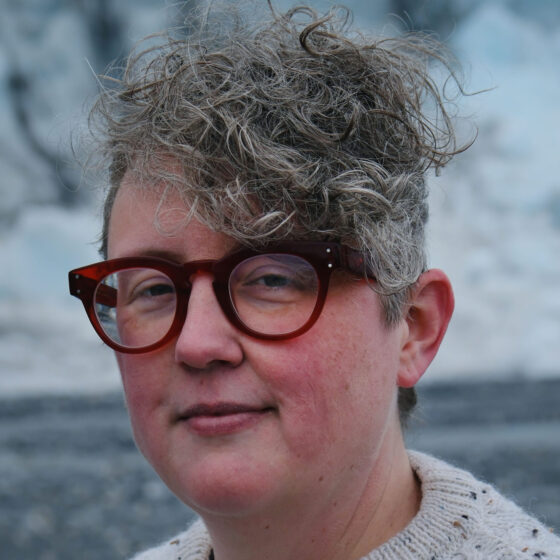 A nonbinary person with gray curly hair on one side of their head. They are wearing red glasses and a cream-colored knit sweater