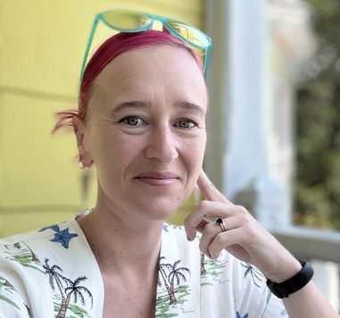 A white woman with a pink bandanna and light-blue sunglasses on her head. She's wearing a white shirt with black palm trees on it and a watch. Her left hand is touching her cheek, and there's yellow siding behind her.