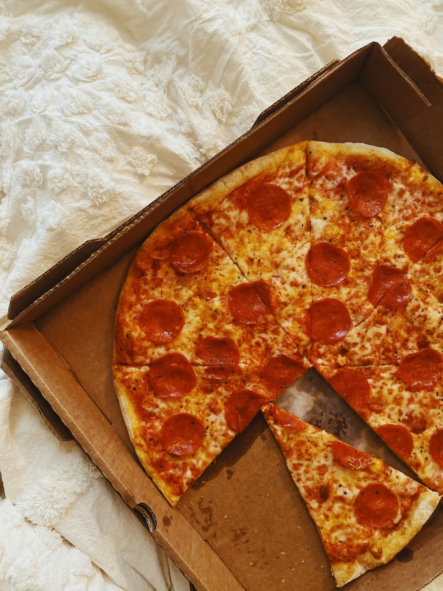 A pepperoni pizza with one slice removed and another turned slightly, in a cardboard box, which is on a white linen