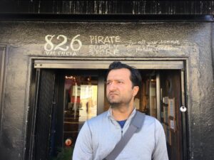 Jaswinder Bolina, a Desi American with dark hair and slight stubble, looking to the left. He's in front of a door that reads "826 Valencia. Pirate Supply Store."