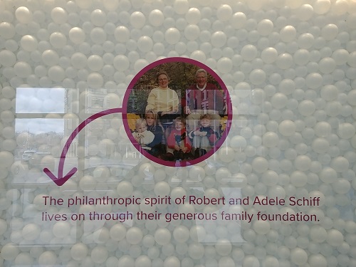 A circle in the middle of a clear wall with bubble shapes behind it. Two grandparents sit with their four grandchildren below them. "The philanthropic spirit of Robert and Adele Schiff lives on through their generous family foundation."