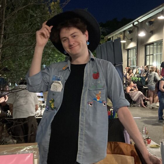Georgia Lavigne, a white person wearing a denim shirt over a black tee, many enamel pins, and a black brimmed hat, tips her hat at the camera. She is outside in a nighttime bookfair setting.