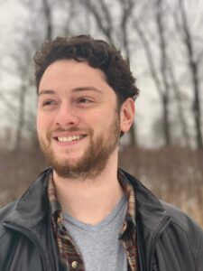 Author Carl Lavigne stands in front of trees in early winter. He has curly brown hair, brown eyes, and a short beard. He wears a black jacket over a flannel shirt. 