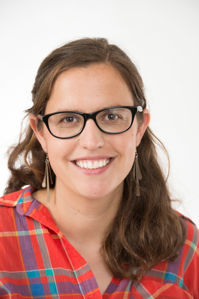 A woman from the shoulders up in red plaid and black glasses smiles directly at the camera