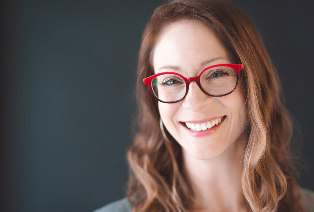 Headshot of a woman smiling at the camera wearing red glasses against a gray background