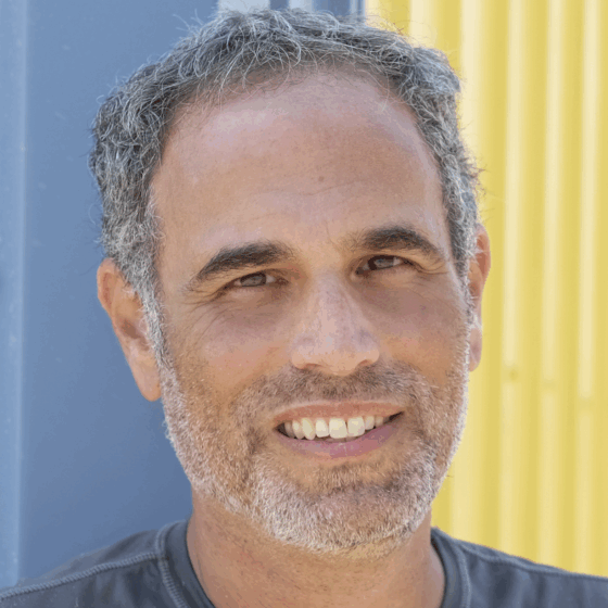A white man with gray hair and trim beard. He's in front of a blue and yellow structure and is wearing a blue-green shirt