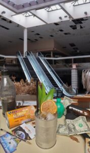 An abandoned mall with a broken escalator is in the background. In the foreground, a Freedom Rampage cocktail in an old soup can sits next to dollar bills and garbage. 