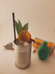 A Freedom Rampage cocktail, decorated with an orange slice and leaves, sits at the center of the image. To the left and right, small plastic tigers circle it. A bottle of lime juice and graffitied can of juice sit to the right. 