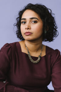 Author SJ Sindu, who has short, curly dark hair, wears a purple shirt and chunky necklace. 