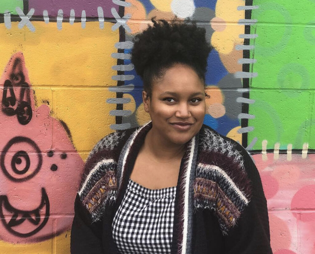 A headshot of a woman smiling with her mouth closed standing in front of a brick wall covered in bright-colored graffiti