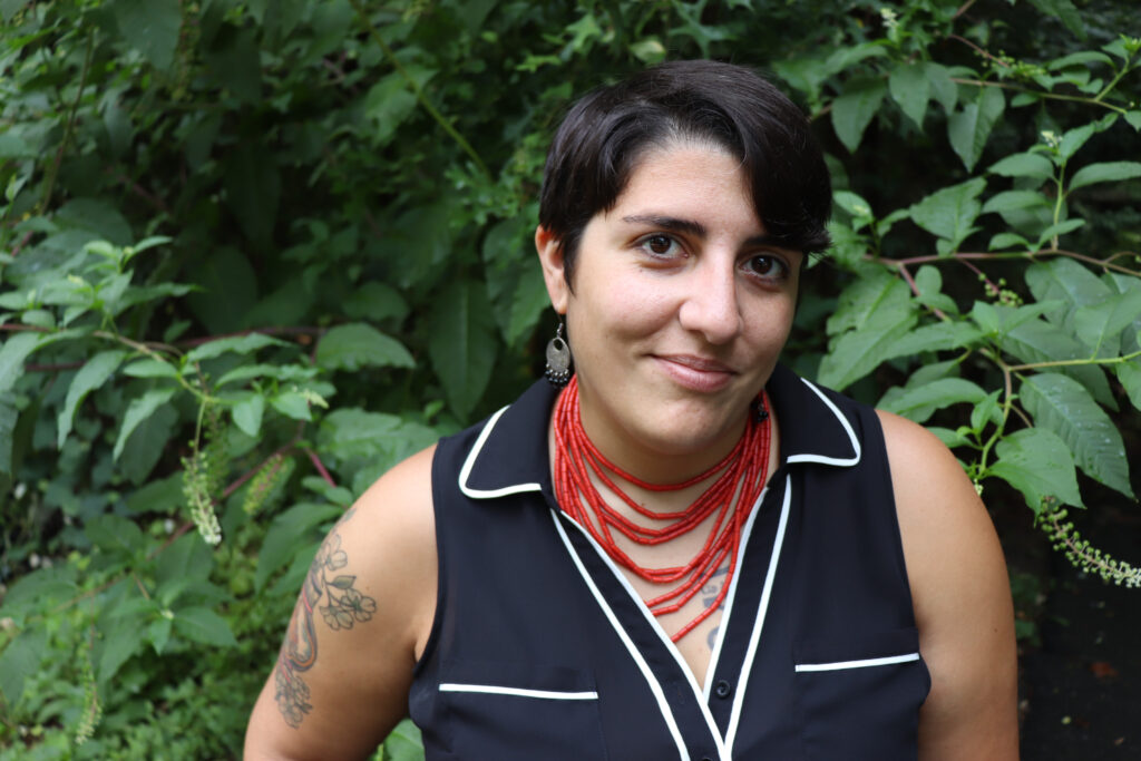 a short-haired woman with a coral bead necklaces and upper arm tattoo stares at the camera