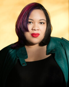 Author Danielle Evans wearing a blue-green jacket over black shirt and staring directly at the camera, with a cream-colored backdrop