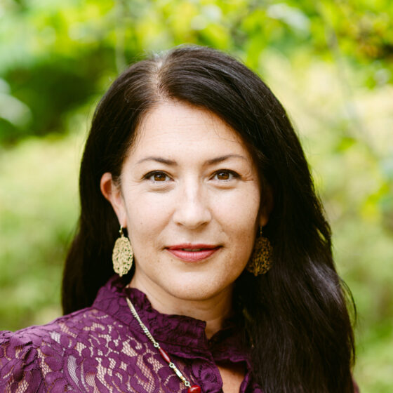 A Latina woman with long dark hair. She's wearing dangling gold leaf earrings and a textured purple blouse, and standing outside