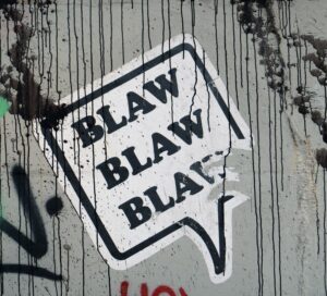 A gray wall covered in black spray paint and dripping black paint. At the center, a sticker reading "Blaw Blaw Blaw" is stuck at a diagonal with a piece missing. 