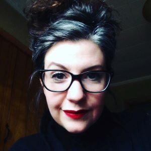 Author Kelly Fig Smith looks directly into the camera. Her dark hair is in a messy bun, and she wears glasses and red lipstick. 