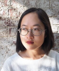 Anni Li, in a white shirt with small dots across the shoulders, looks to the left of the photo, with a red-and-white brick wall behind her.