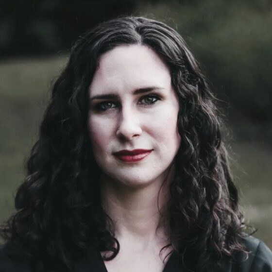 A dark photo of a woman with long, dark, curly hair and dark eyes, smiling. She is standing outside and wearing a black shirt.