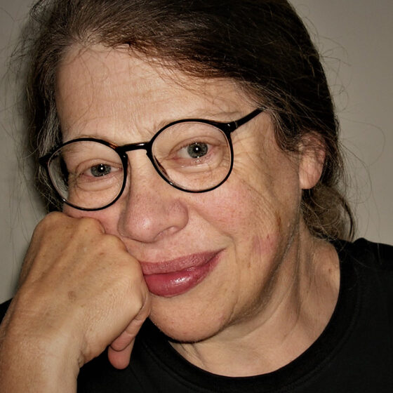 A woman with long, straight, brown hair and brown eyes, smiling. She leans her face on her right fist, wearing a black shirt and round-square glasses