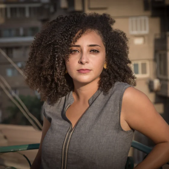 A woman with shoulder-length, brown, curly hair and brown eyes. She stands outside in front of some concrete buildings wearing a gray tank top.