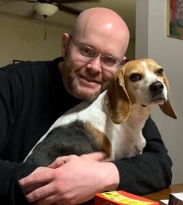 Steven D. Schroeder, wearing glasses and a dark sweater and holding a beagle
