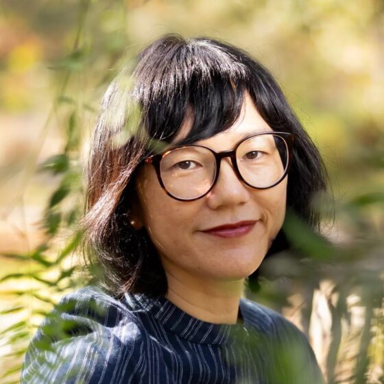 An Asian American woman with short dark hair and wide glasses. She's wearing a blue striped button-up blouse and is sitting on a bench, with greenery slightly between her and the camera.