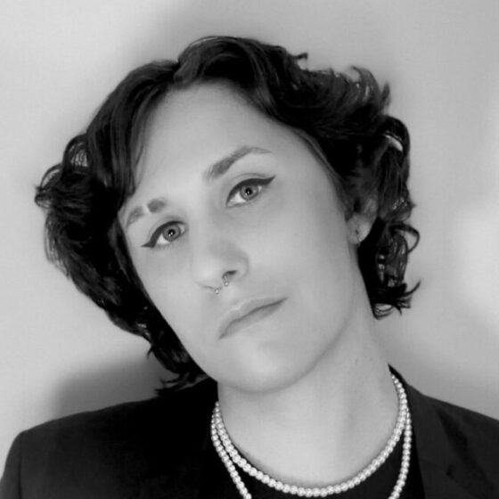Black and white photo of a trans woman with short dark curly hair, leaning her head toward the left side of the image. She's wearing a dark jacket over a darker shirt and also a string of pearls.
