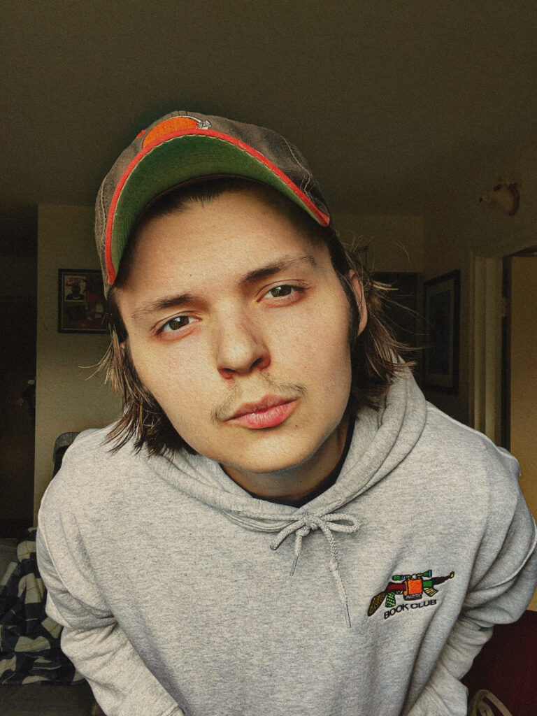 Matt Mitchell wears a grey sweatshirt and a orange and green ball cap as he leans in towards the camera.