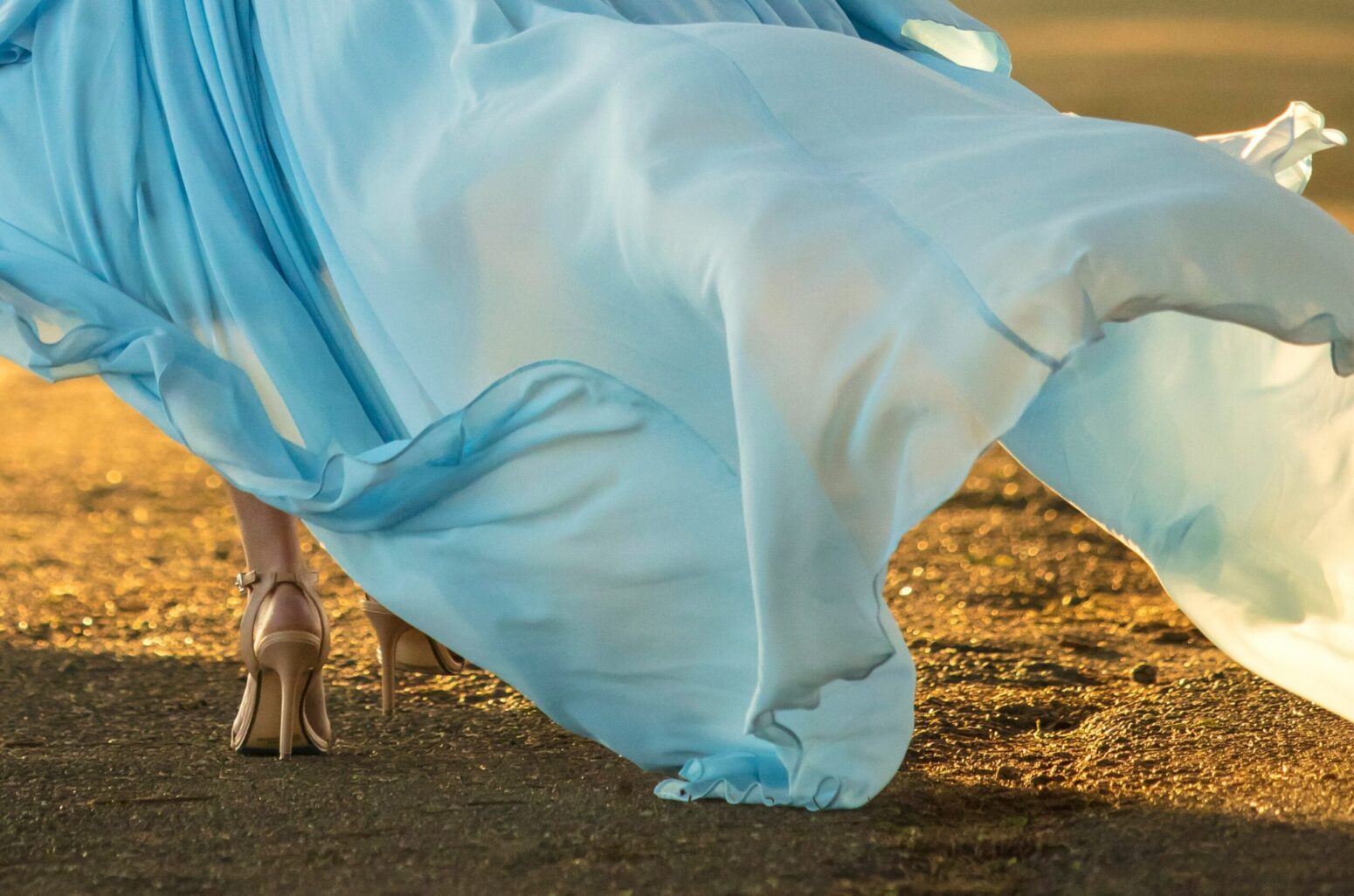 A woman from the knees down, wearing heels and a long, flowing, blue dress and walking on a dirt path