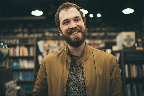 Connor Yeck, with a beard and brown hair, smiling. He's wearing a tan jacket and black-and-white marbled shirt. In the background are bookcases, out of focus.