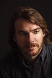 Robert Wood Lynn headshot. He has a short beard and collar-length hair. He wears a black shirt and poses against a black background.