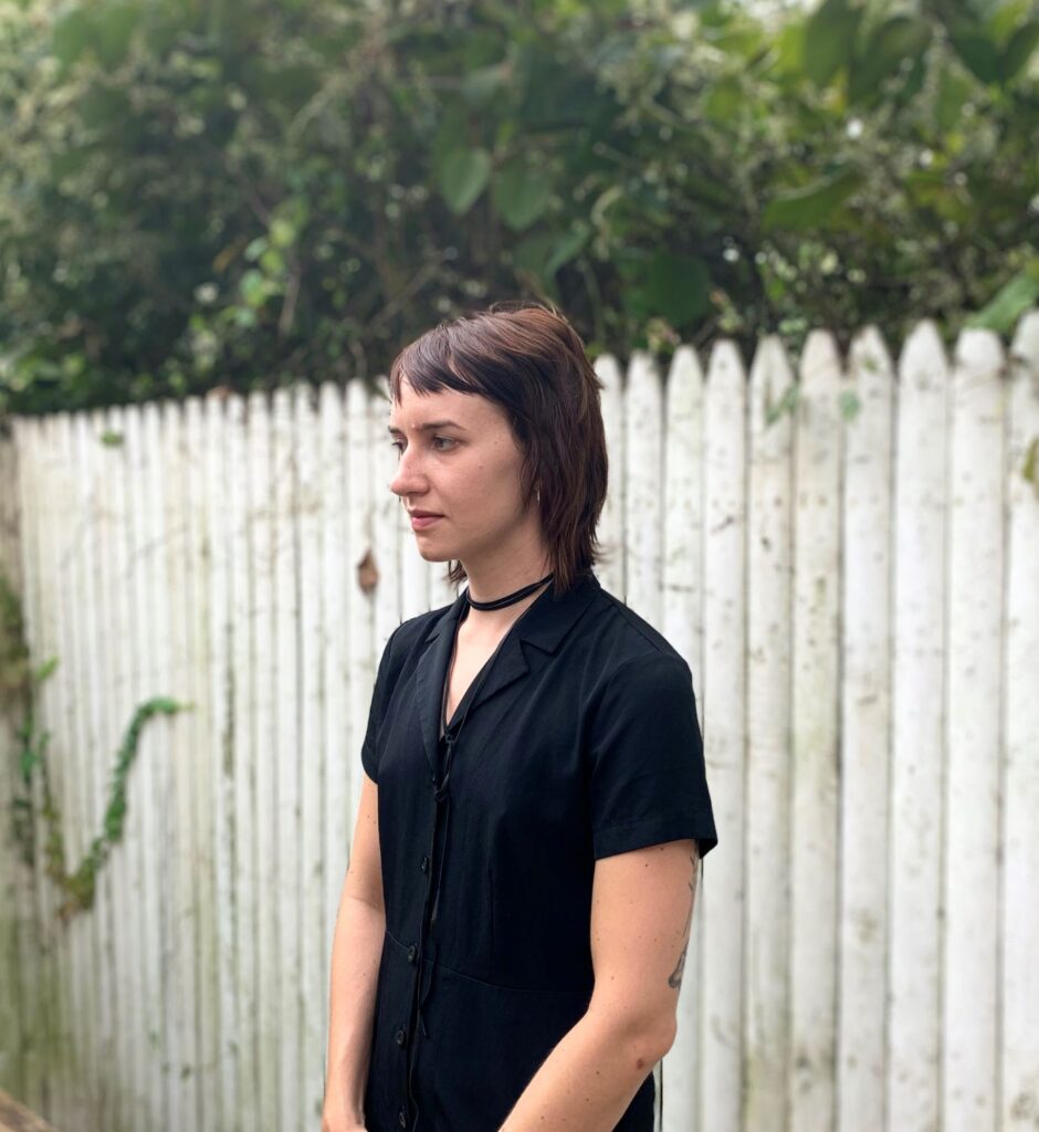 Photo of author wearing a black top and a choker, standing in front of a white fence and looking to the side. 