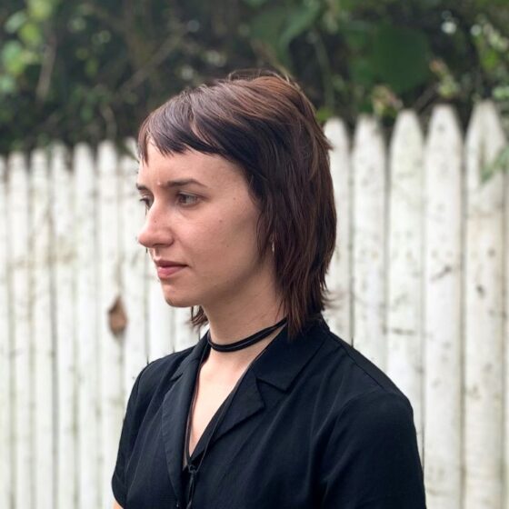 A white woman with short dark hair and a choker-style black necklace and black blouse. She is turned to the side and stands in front of a white picket fence and greenery, without expression.