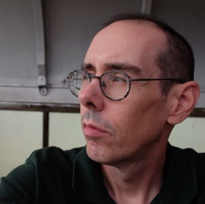 Photo of author looking to the left, wearing round glasses and a dark green shirt. 