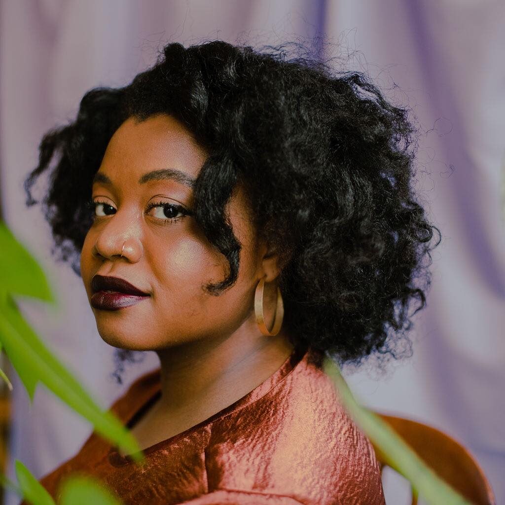 Photo of author wearing a dark orange shirt and hoops, against a purple background with plants