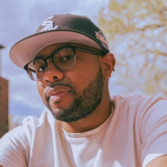 A man with a short, black beard and brown eyes. He wears a white t-shirt, square-shaped glasses, and a black hat
